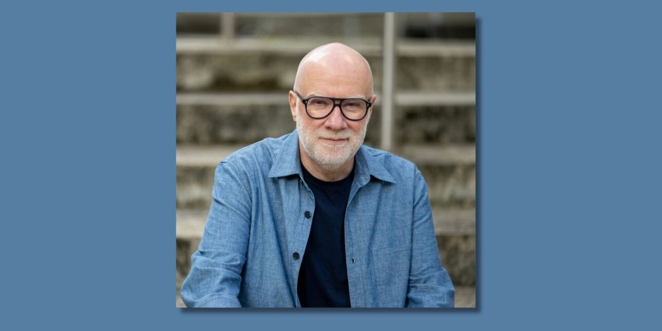 A colour portrait of John Spencer Williams, a white man with grey beard and glasses, who is wearing a blue shirt.